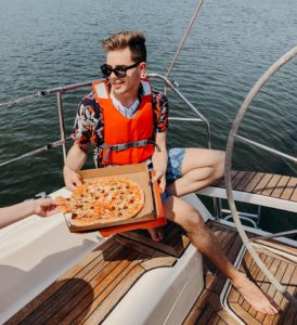 Man on boat with a lifejacket, sunglasses, and pizza.